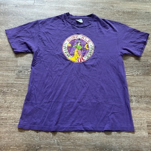 May include: Purple t-shirt with a circular graphic featuring a witch-like figure in green and yellow, with the text "RUBY ALE" in a circular design. The graphic is set against a purple and starry background.