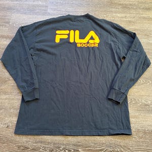 Peut inclure: Un t-shirt &agrave; manches longues gris fonc&eacute; avec le texte jaune "FILA SOCCER" au dos. Le t-shirt a un col rond et des manches longues avec des poignets c&ocirc;tel&eacute;s. Le t-shirt est fait d'une mati&egrave;re douce et confortable.