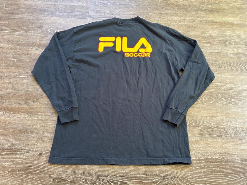 Peut inclure: Un t-shirt &agrave; manches longues gris fonc&eacute; avec le texte jaune "FILA SOCCER" au dos. Le t-shirt a un col rond et des manches longues avec des poignets c&ocirc;tel&eacute;s. Le t-shirt est fait d'une mati&egrave;re douce et confortable.