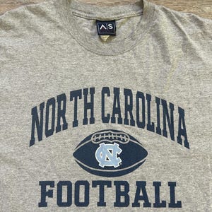 Może przedstawiać: Szara koszulka z napisem "NORTH CAROLINA" wygiętym łukiem u g&oacute;ry i "FOOTBALL" pod grafiką piłki nożnej. Grafika piłki nożnej ma jasnoniebieskie logo "NC" pośrodku.