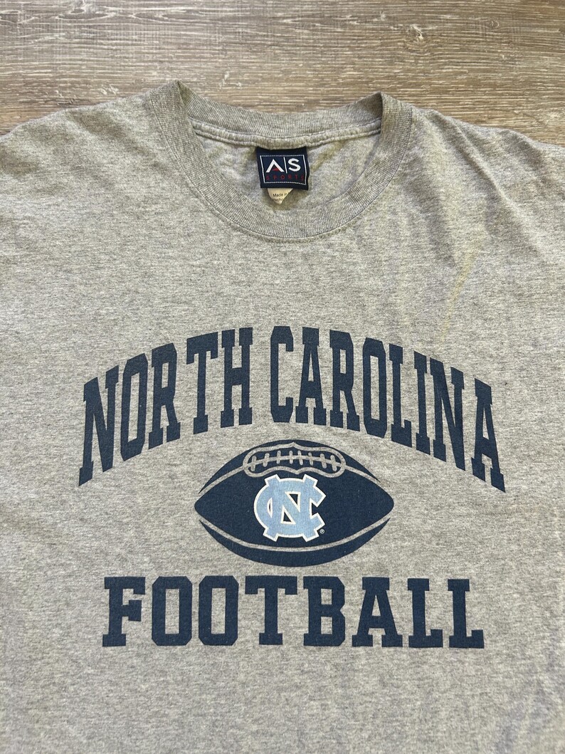 Może przedstawiać: Szara koszulka z napisem "NORTH CAROLINA" wygiętym łukiem u g&oacute;ry i "FOOTBALL" pod grafiką piłki nożnej. Grafika piłki nożnej ma jasnoniebieskie logo "NC" pośrodku.