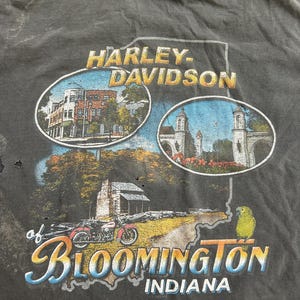 May include: Dark gray Harley-Davidson t-shirt featuring a graphic of Bloomington, Indiana. The design includes illustrations of buildings, a motorcycle, and a parrot. The text reads "Harley-Davidson of Bloomington Indiana."