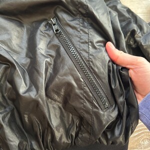 Pode incluir: Casaco preto de nylon com um bolso com z&iacute;per e bainha canelada. O casaco tem um aspeto brilhante e ligeiramente enrugado. O z&iacute;per &eacute; prateado.