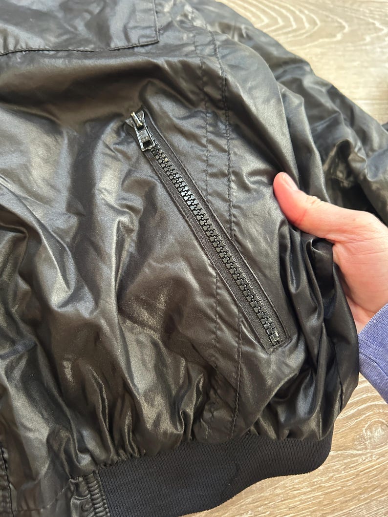 Pode incluir: Casaco preto de nylon com um bolso com z&iacute;per e bainha canelada. O casaco tem um aspeto brilhante e ligeiramente enrugado. O z&iacute;per &eacute; prateado.