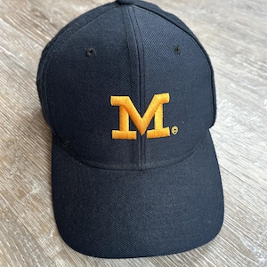 May include: A navy blue baseball cap with a curved brim. The cap features an embroidered gold letter "M" on the front. The cap is made of a durable fabric and has a structured fit.