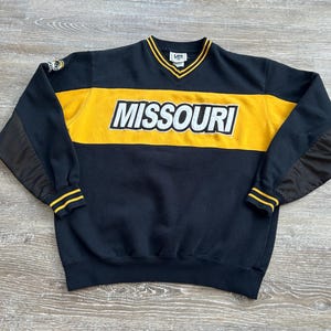 May include: A black and gold long-sleeve sweatshirt featuring the word MISSOURI in white block letters across the chest. The garment has a V-neck collar with yellow and black stripes, mesh panels on the sleeves, and a Lee brand label at the neck.