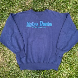 May include: A navy blue long-sleeve crewneck sweatshirt featuring the text Notre Dame and Fighting Irish embroidered in light blue thread across the chest. The garment has ribbed cuffs and a ribbed hem, with a V-shaped stitch detail at the collar.