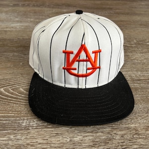 May include: A white and black pinstripe baseball cap with a black brim. The cap features an orange embroidered logo on the front. The cap is a sports-themed accessory.