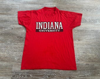 Könnte beinhalten: Ein rotes T-Shirt mit dem Text "INDIANA UNIVERSITY" in weißen Blockbuchstaben mit schwarzem Rand. Der Text ist mittig auf der Brust platziert. Das Shirt besteht aus einem weichen Material.