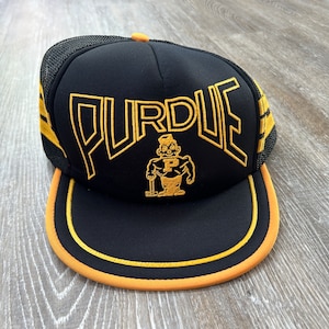 May include: Black and gold trucker hat with the word "PURDUE" in a yellow outline and a cartoon mascot wearing a yellow shirt with a "P" on it.