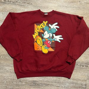 May include: A maroon sweatshirt featuring a graphic of Mickey Mouse. The cartoon character is depicted in a dynamic pose with his name in yellow letters. The design includes orange, yellow, red, and white accents.