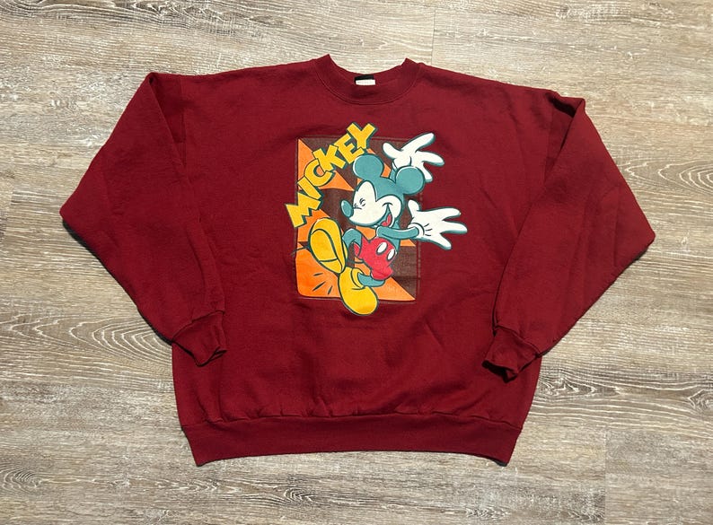 May include: A maroon sweatshirt featuring a graphic of Mickey Mouse. The cartoon character is depicted in a dynamic pose with his name in yellow letters. The design includes orange, yellow, red, and white accents.