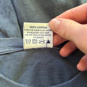 Peut inclure: Gros plan sur une &eacute;tiquette de v&ecirc;tement avec le texte "100% COTON" et des instructions de lavage. L'&eacute;tiquette est attach&eacute;e &agrave; un t-shirt bleu. Les instructions de lavage incluent lavage en machine &agrave; l'eau froide, s&eacute;chage en machine &agrave; basse temp&eacute;rature, ne pas repasser si d&eacute;cor&eacute; et pas de javel.