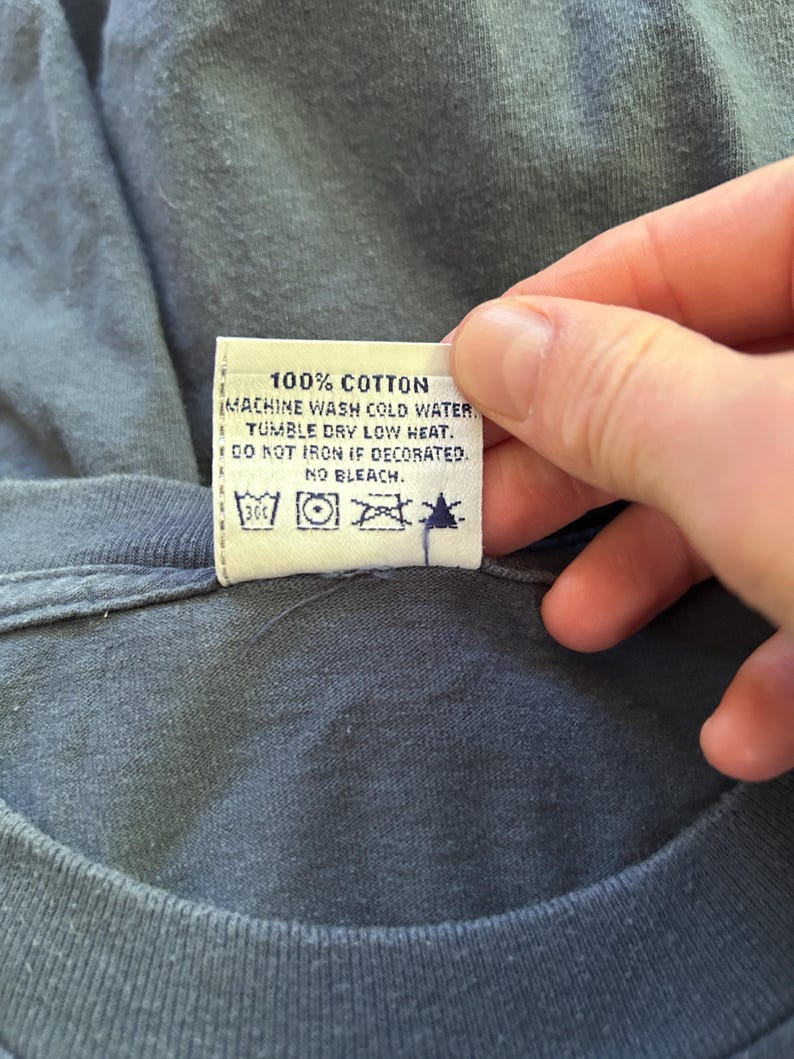 Peut inclure: Gros plan sur une &eacute;tiquette de v&ecirc;tement avec le texte "100% COTON" et des instructions de lavage. L'&eacute;tiquette est attach&eacute;e &agrave; un t-shirt bleu. Les instructions de lavage incluent lavage en machine &agrave; l'eau froide, s&eacute;chage en machine &agrave; basse temp&eacute;rature, ne pas repasser si d&eacute;cor&eacute; et pas de javel.