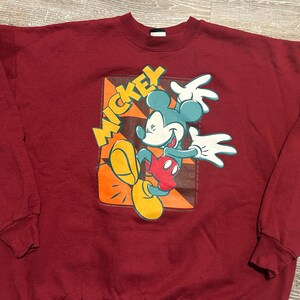 May include: Maroon sweatshirt featuring a colorful graphic of Mickey Mouse. The design includes Mickey in a dynamic pose with yellow, orange, red, and blue accents, and the word "Mickey" in yellow. The sweatshirt has a crew neck and long sleeves.
