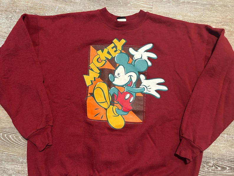May include: Maroon sweatshirt featuring a colorful graphic of Mickey Mouse. The design includes Mickey in a dynamic pose with yellow, orange, red, and blue accents, and the word "Mickey" in yellow. The sweatshirt has a crew neck and long sleeves.