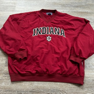 May include: A red pullover windbreaker with a V-neck collar. The word "INDIANA" is printed across the chest in white block letters with a black outline. Below the word is a white and black logo.