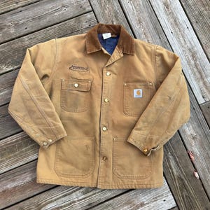 May include: A tan Carhartt work jacket with a brown corduroy collar and a blue denim lining. The jacket has four front pockets, gold-colored buttons, and a small patch that says "Ameritech".