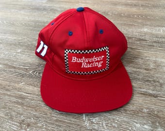 May include: A red baseball cap with a curved brim. The cap features the Budweiser Racing logo on the front, a white number 77 on the side, and blue accents. The cap is made of a durable fabric.