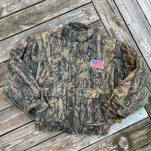 May include: Camouflage jacket with an American flag patch on the chest. The jacket features a button-up front, two chest pockets, and a high collar. The pattern is a mix of green, brown, and tan, designed to blend into a wooded environment.