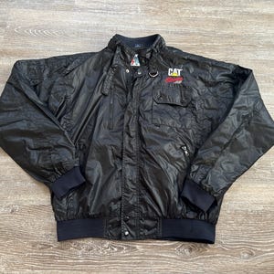 Pode incluir: Casaco de nylon preto com gola alta, v&aacute;rios bolsos e fecho de correr frontal. O casaco tem um log&oacute;tipo CAT no peito e punhos e bainha canelados. O casaco est&aacute; estendido sobre uma superf&iacute;cie de madeira.