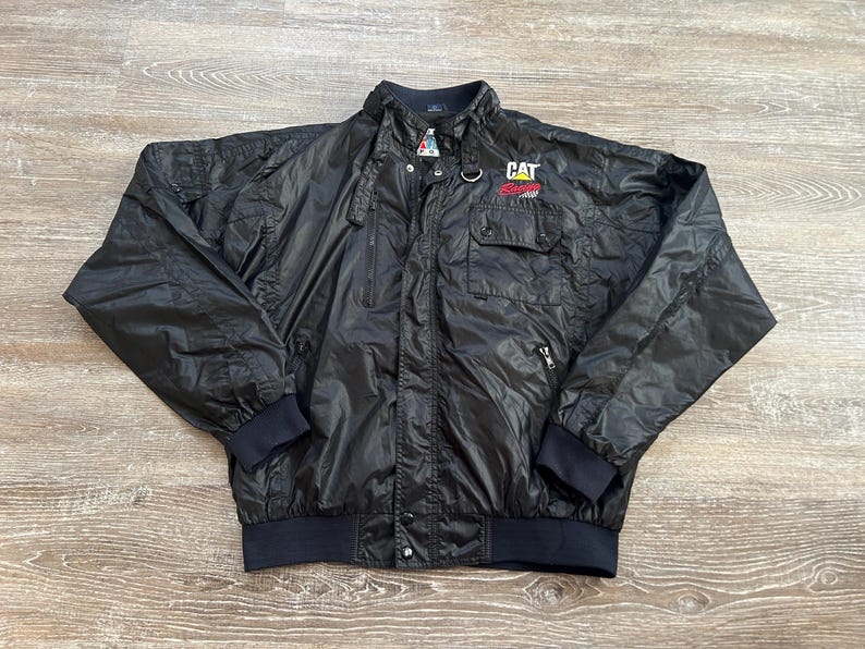 Pode incluir: Casaco de nylon preto com gola alta, v&aacute;rios bolsos e fecho de correr frontal. O casaco tem um log&oacute;tipo CAT no peito e punhos e bainha canelados. O casaco est&aacute; estendido sobre uma superf&iacute;cie de madeira.