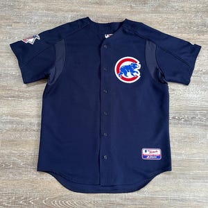 May include: Navy blue baseball jersey featuring the Chicago Cubs logo on the chest. The jersey has a button-up front, short sleeves, and a patch on the left sleeve. The Cubs logo is a blue bear inside a red circle.