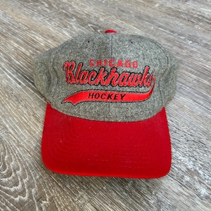 May include: A gray and red Chicago Blackhawks hockey cap. The cap features the team name in red script with a red banner that reads "HOCKEY". The brim is red, and the cap is made of a wool-like material.