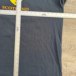 Peut inclure: T-shirt bleu fonc&eacute; avec un embl&egrave;me &eacute;cossais jaune et rouge. Le mot "SCOTLAND" est imprim&eacute; en or. Le t-shirt est mesur&eacute; avec une r&egrave;gle. Le t-shirt mesure environ 76 cm de long.