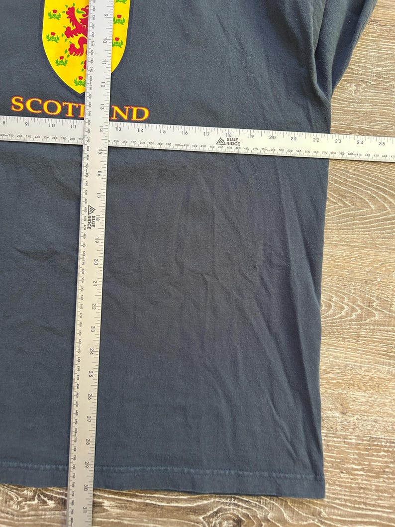 Peut inclure: T-shirt bleu fonc&eacute; avec un embl&egrave;me &eacute;cossais jaune et rouge. Le mot "SCOTLAND" est imprim&eacute; en or. Le t-shirt est mesur&eacute; avec une r&egrave;gle. Le t-shirt mesure environ 76 cm de long.