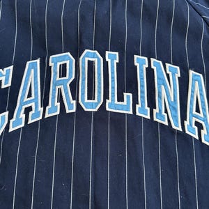 Pu&ograve; includere: Tessuto blu navy con strisce verticali bianche. La parola "CAROLINA" &egrave; arcuata sul tessuto in lettere azzurre con contorni bianchi. Il tessuto sembra far parte di un capo d'abbigliamento.