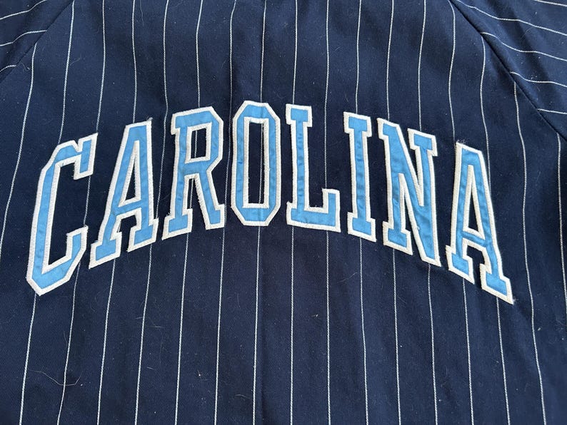 Pu&ograve; includere: Tessuto blu navy con strisce verticali bianche. La parola "CAROLINA" &egrave; arcuata sul tessuto in lettere azzurre con contorni bianchi. Il tessuto sembra far parte di un capo d'abbigliamento.