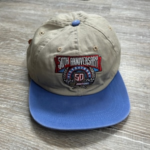 May include: A beige and blue baseball cap with a 50th Anniversary Nascar logo embroidered on the front. The logo features a red, white, and blue shield with the number 50 inside.