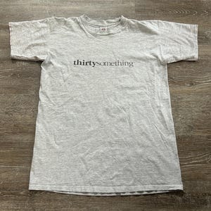 May include: A light gray t-shirt with the word "thirtysomething" printed in black across the chest. The shirt has short sleeves and a crew neck. The fabric appears to be a soft cotton blend.