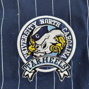 Pu&ograve; includere: Toppa ricamata con il logo dei Tar Heels dell'Universit&agrave; della Carolina del Nord. La toppa &egrave; circolare, con il nome della squadra e una mascotte di ariete. Lo sfondo &egrave; blu navy con strisce verticali bianche.