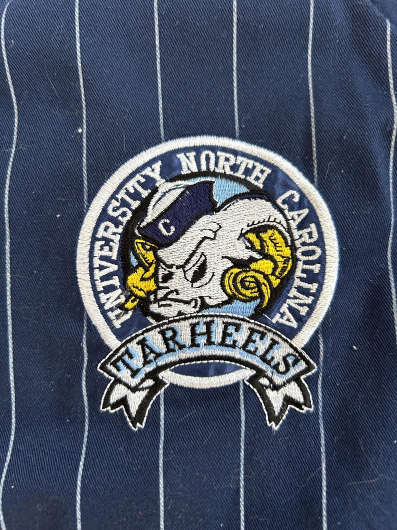Pu&ograve; includere: Toppa ricamata con il logo dei Tar Heels dell'Universit&agrave; della Carolina del Nord. La toppa &egrave; circolare, con il nome della squadra e una mascotte di ariete. Lo sfondo &egrave; blu navy con strisce verticali bianche.