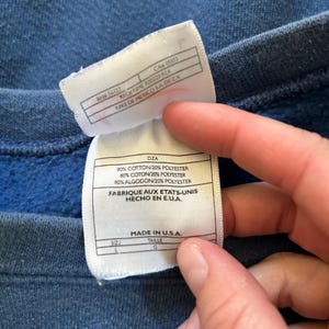 May include: A white tag with black text detailing the fabric content and origin of the garment. The tag reads "80% Cotton/20% Polyester, 80% Coton/20% Polyester, 80% Algodon/20% Polyester, Fabrique aux Etats-Unis, Hecho en E.U.A., Made in U.S.A.". The tag also indicates the size as "L" and the taille as "G".