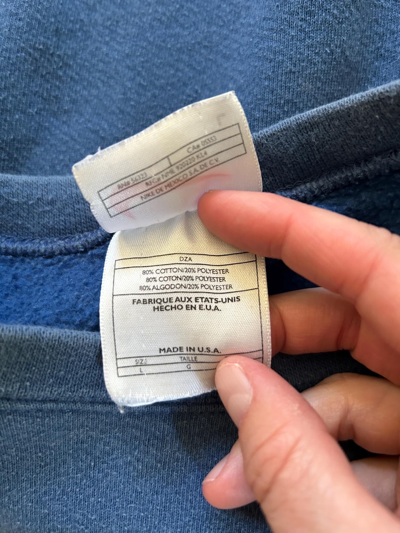 May include: A white tag with black text detailing the fabric content and origin of the garment. The tag reads "80% Cotton/20% Polyester, 80% Coton/20% Polyester, 80% Algodon/20% Polyester, Fabrique aux Etats-Unis, Hecho en E.U.A., Made in U.S.A.". The tag also indicates the size as "L" and the taille as "G".