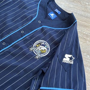 Pu&ograve; includere: Maglia da baseball blu navy con gessati bianchi, rifiniture azzurre e logo Starter. La maglia presenta uno stemma dell'Universit&agrave; della Carolina del Nord Tar Heels sul petto.