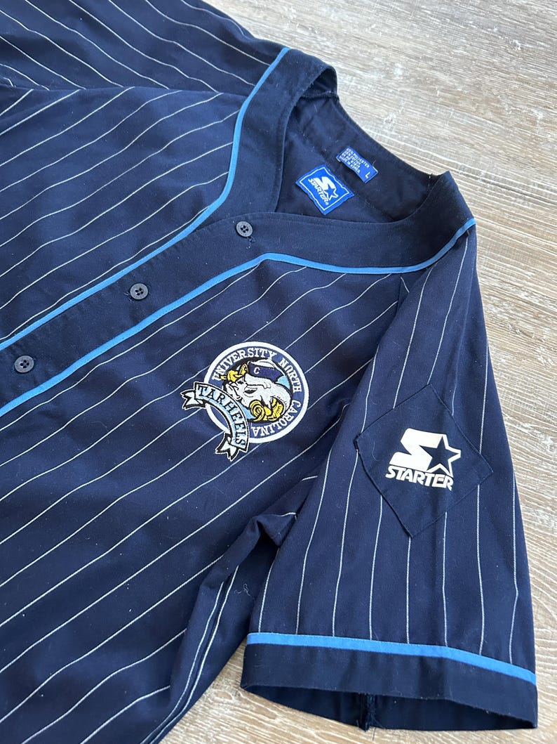 Pu&ograve; includere: Maglia da baseball blu navy con gessati bianchi, rifiniture azzurre e logo Starter. La maglia presenta uno stemma dell'Universit&agrave; della Carolina del Nord Tar Heels sul petto.