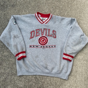 May include: A heather gray long-sleeve sweatshirt featuring a V-neck collar with red and white stripes. The front displays the text DEVILS in red with black outlines, a circular logo, and the text NEW JERSEY. Red and white stripes accent the cuffs.