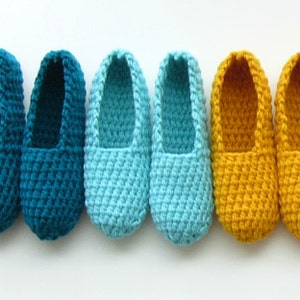 Può includere: Cinque paia di pantofole all'uncinetto in tonalità di blu-verde, blu e giallo. Le pantofole sono allineate in una fila dal più scuro al più chiaro.