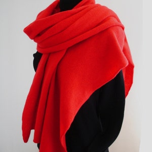 Pode incluir: Um cachecol vermelho vibrante drapeado sobre um top preto. O cachecol é feito de um material macio e texturizado e é enrolado ao redor do pescoço e dos ombros. A cor brilhante do cachecol contrasta com as roupas escuras.