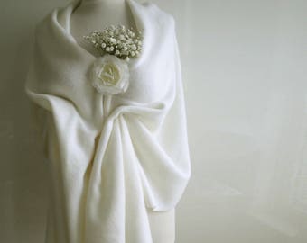 May include: A cream-colored shawl draped over a mannequin, adorned with a white rose and small white flowers. The shawl is made of a soft, textured fabric, creating elegant folds. The overall aesthetic is delicate and refined.