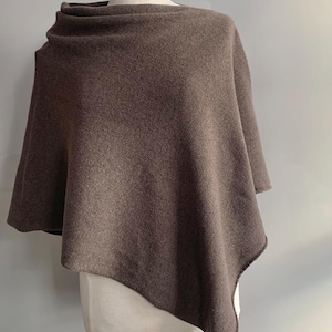 Pode incluir: Um poncho marrom com gola alta, exibido em um manequim. O poncho é feito de um tecido macio e texturizado. A peça tem uma bainha assimétrica e fluida e drapeja sobre os ombros.