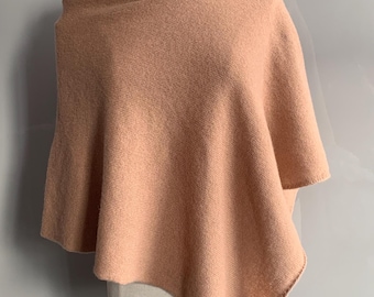 May include: A peach-colored poncho made of a soft, textured fabric. The poncho is draped over a mannequin, showcasing its asymmetrical hemline and boat neck design. The garment appears warm and comfortable, suitable for cooler weather.