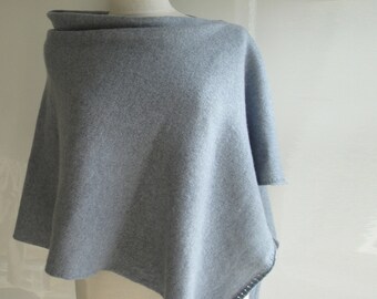 May include: A light blue poncho made of soft fabric. The poncho has a wide, folded collar and a slightly asymmetrical hemline. The edges are finished with a dark gray stitch detail. The garment is displayed on a mannequin.