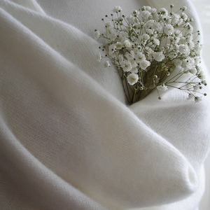 Puede incluir: Un chal de punto blanco crema, con un ramo de peque&ntilde;as flores blancas metido en sus pliegues. El chal est&aacute; hecho de un material suave y ligero, creando un aspecto acogedor y elegante. Las flores a&ntilde;aden un toque de belleza natural a la prenda.