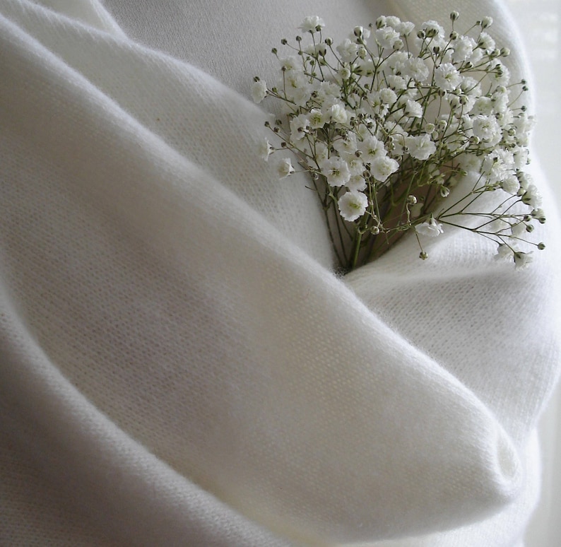 Puede incluir: Un chal de punto blanco crema, con un ramo de peque&ntilde;as flores blancas metido en sus pliegues. El chal est&aacute; hecho de un material suave y ligero, creando un aspecto acogedor y elegante. Las flores a&ntilde;aden un toque de belleza natural a la prenda.