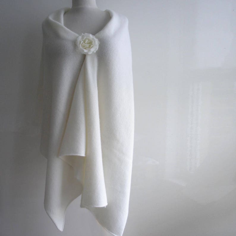 Puede incluir: Un chal blanco drapeado sobre un maniqu&iacute;, asegurado con un broche de flores blanco. El chal tiene una apariencia suave y texturizada y un dise&ntilde;o drapeado. El fondo es una pared blanca lisa.